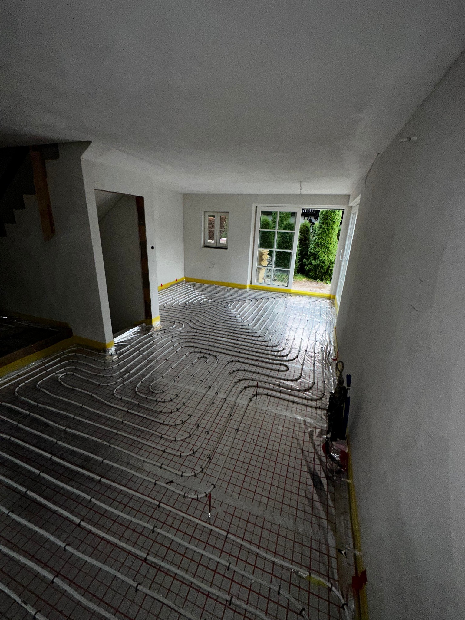 Wnętrze w trakcie instalacji ogrzewania podłogowego: białe rurki układane na siatce z czerwonymi oczkami, widoczne okno z zielenią na zewnątrz, przygotowane do wylania posadzki.