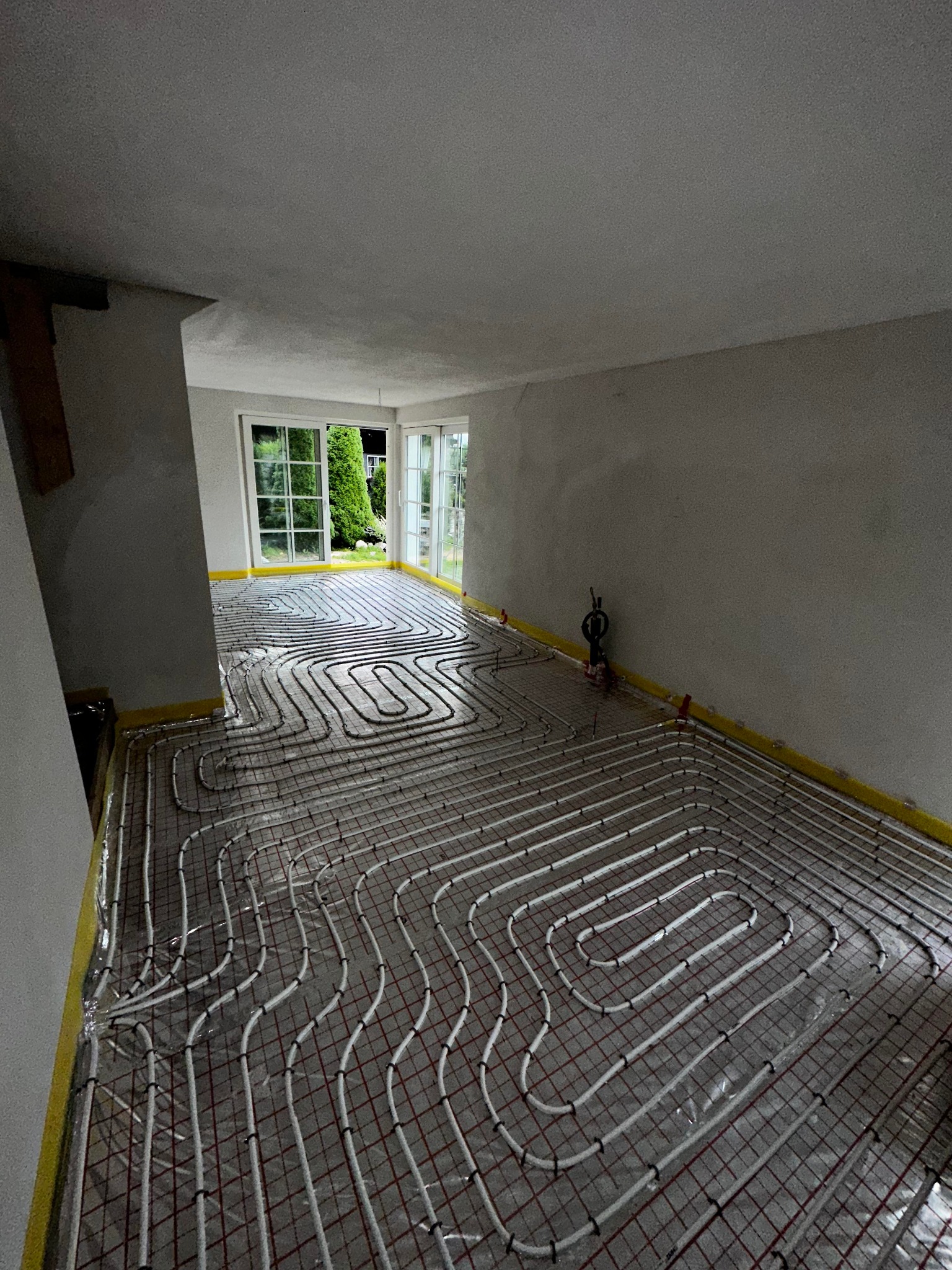 Rozłożona instalacja ogrzewania podłogowego z rurkami na folii, widok na okno z zielenią w tle, przygotowana do zalania wylewką.