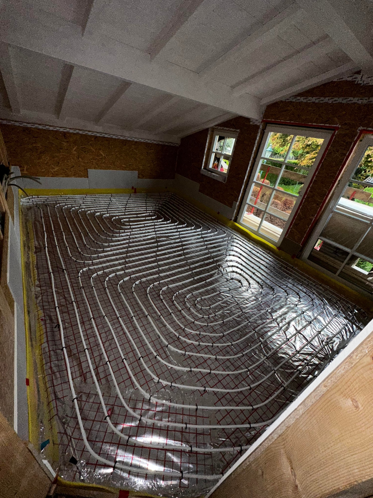 Układanie rur ogrzewania podłogowego na folii odblaskowej w pomieszczeniu z oknami, widok z góry na etap prac instalacyjnych.