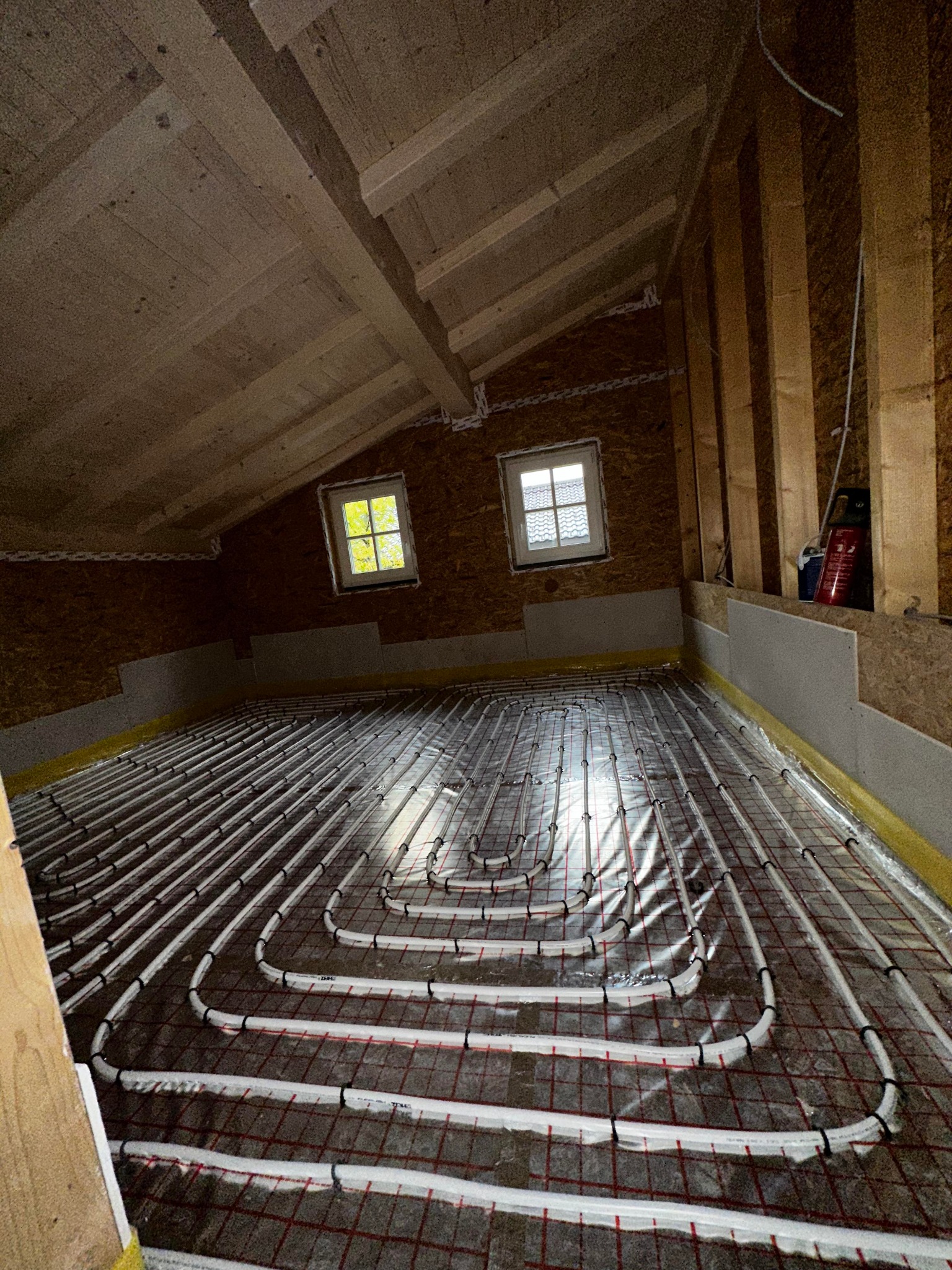 Instalacja ogrzewania podłogowego w nowym budynku na poddaszu, rury grzewcze ułożone na siatce, widoczne okna dachowe i surowa konstrukcja drewniana.