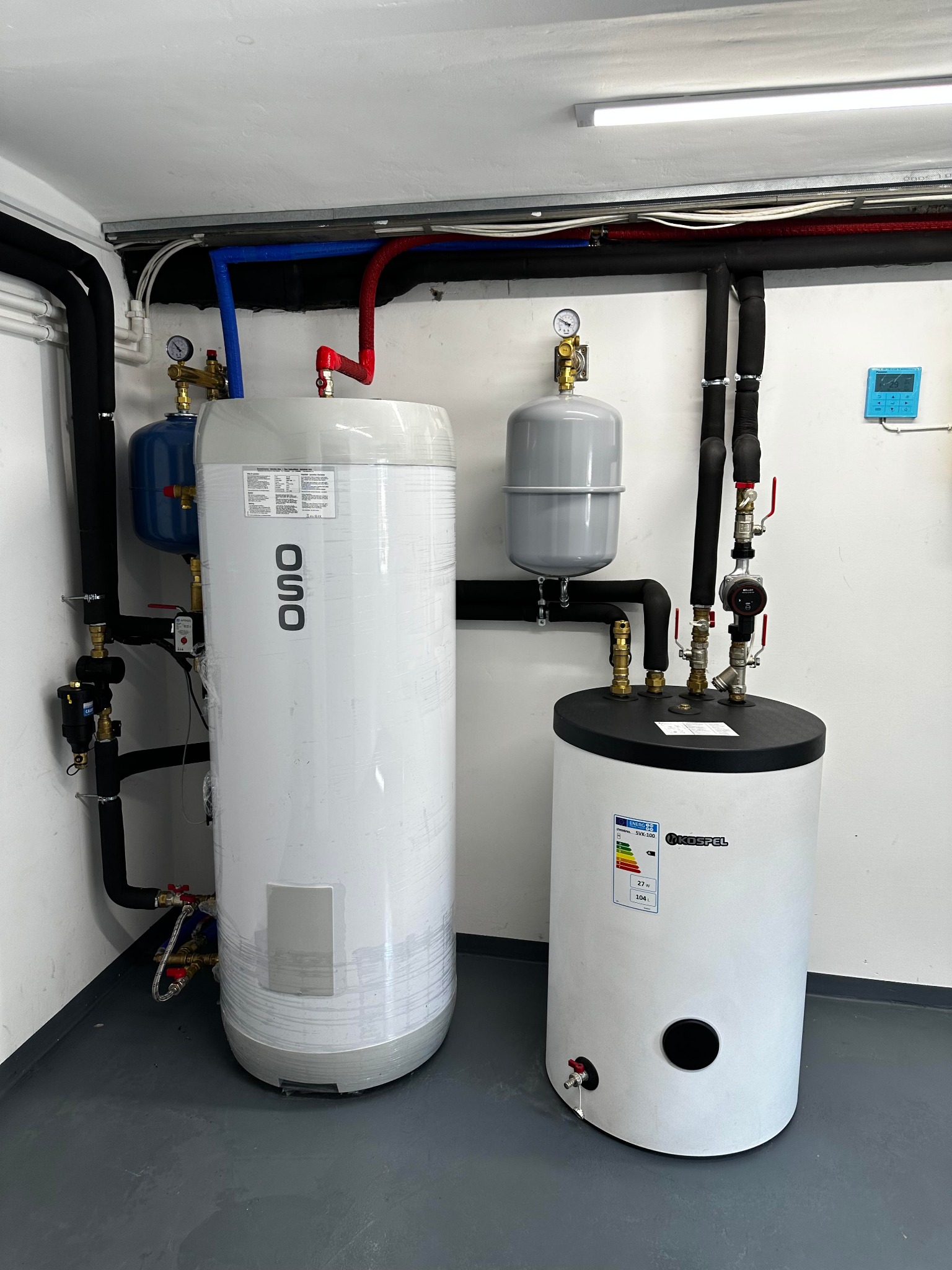 Dwa białe zasobniki ciepłej wody różnych marek (OSO i Kospel) z podłączeniami hydraulicznymi, zaworami i rurami w kotłowni na szarej podłodze.
