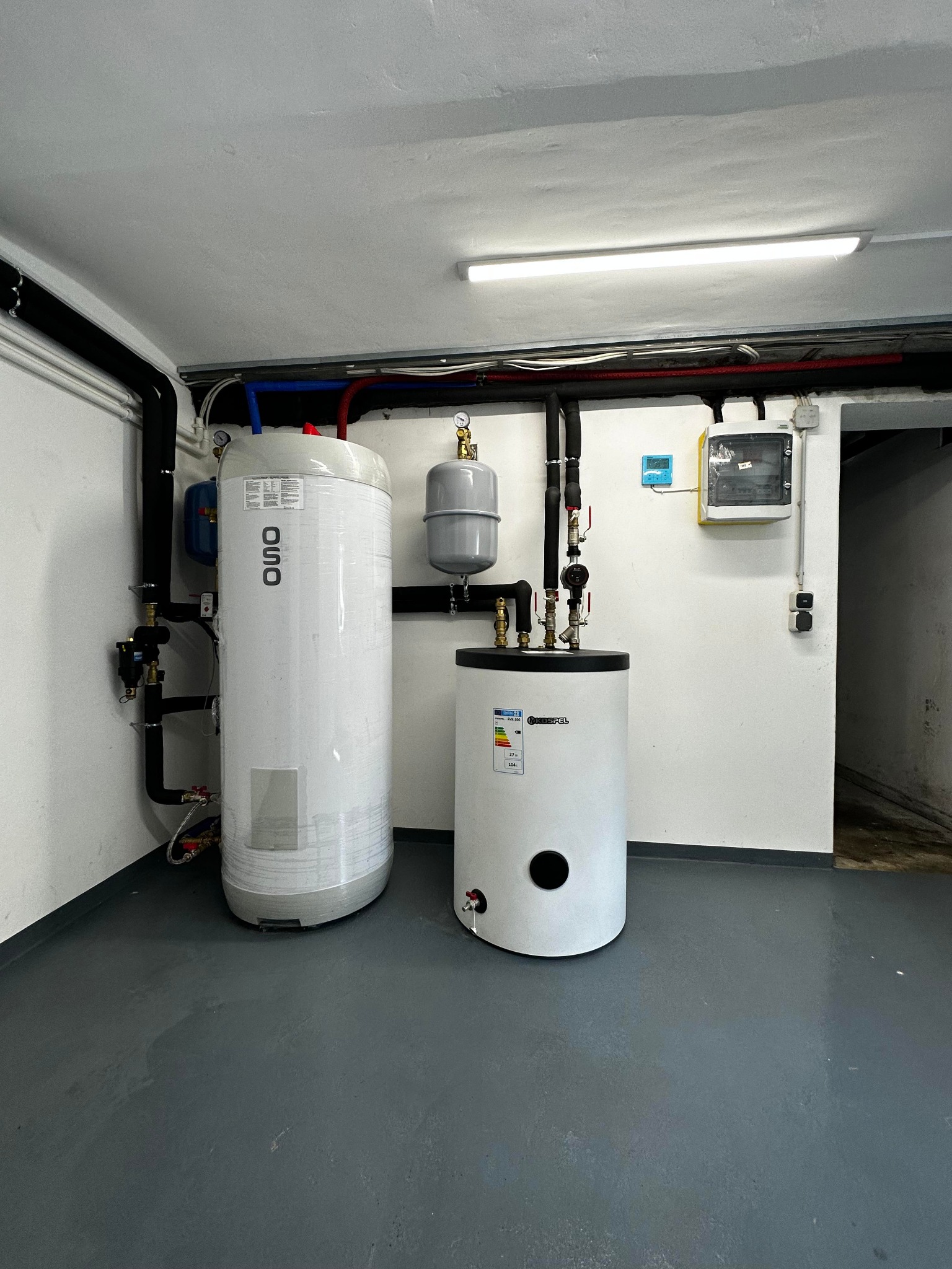 Dwa białe zbiorniki akumulacyjne, jeden marki OSO w folii ochronnej, drugi Kospel, zainstalowane w kotłowni w Pietnej z szarym betonowym podłożem i rurami pod sufitem.