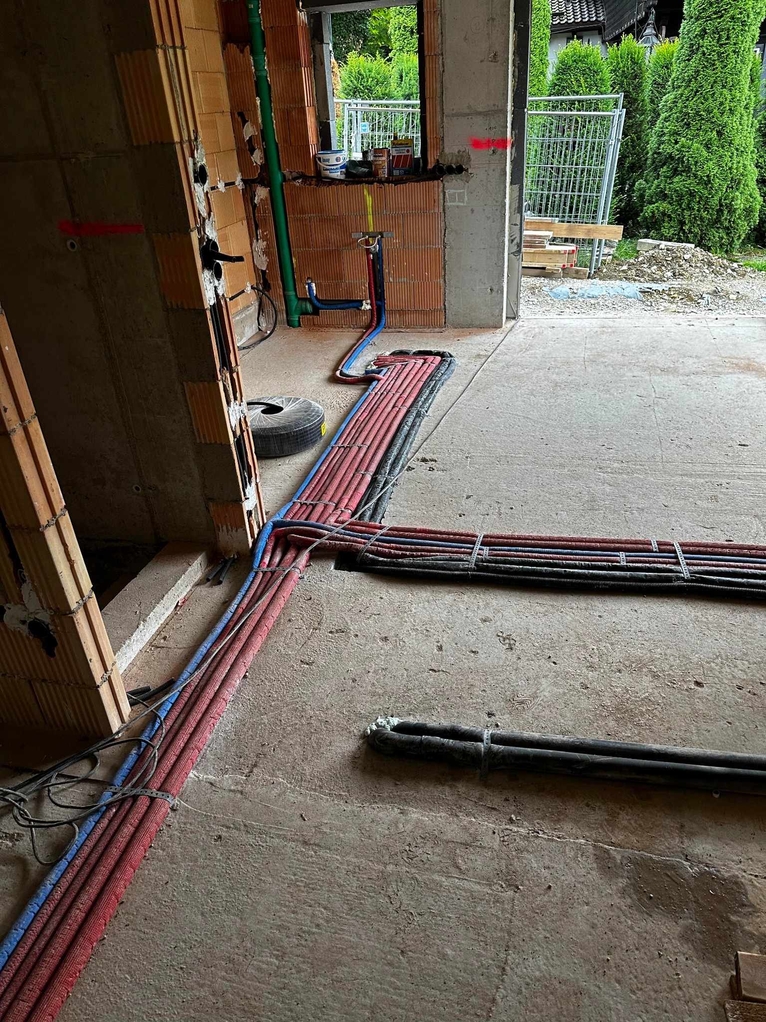 Wnętrze budynku w trakcie instalacji hydraulicznej i grzewczej: liczne rury w kolorach czerwonym, niebieskim i czarnym prowadzone po posadzce, ściany z pustaków ceramicznych z widocznymi...