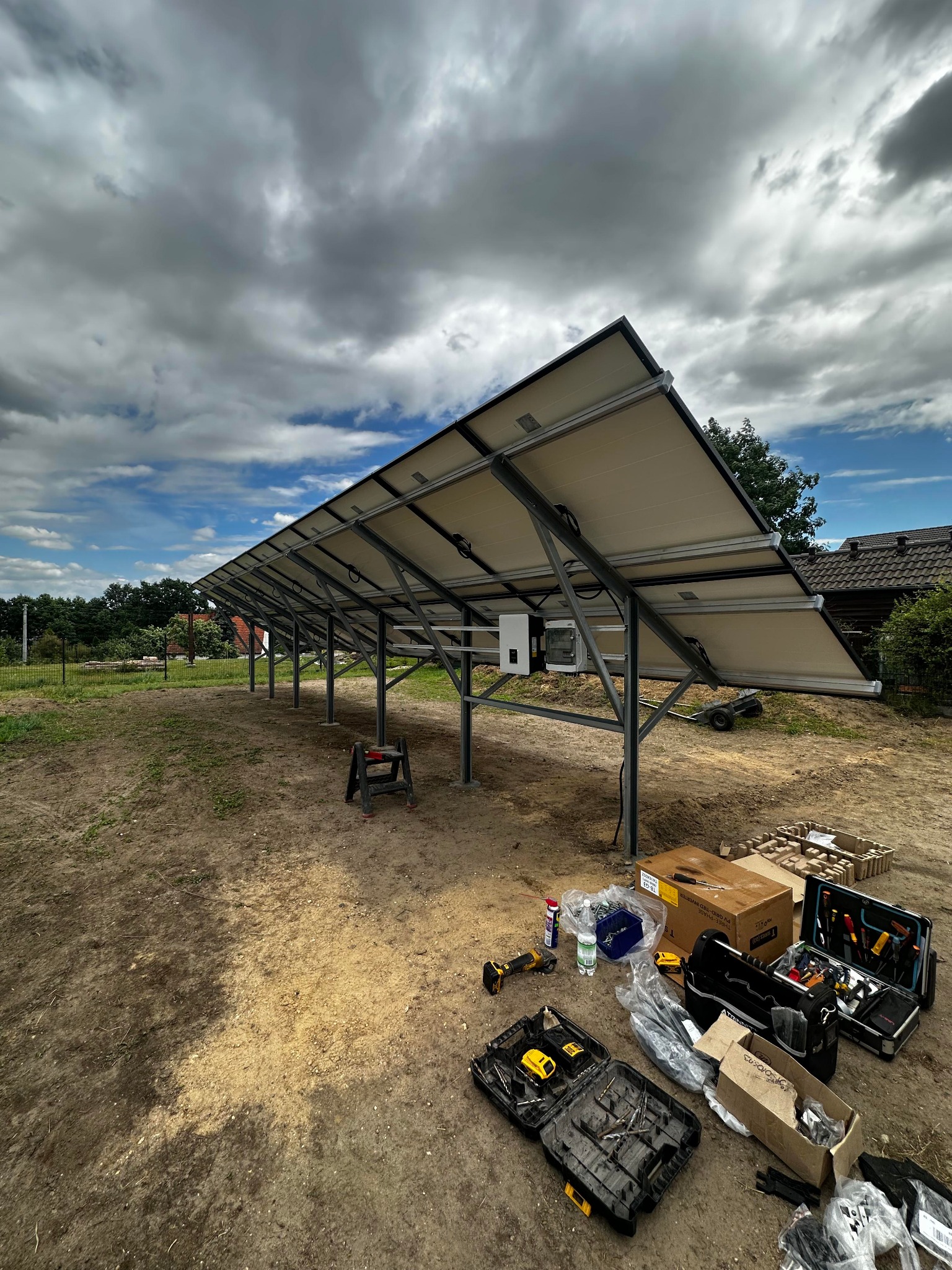 Montaż paneli słonecznych na konstrukcji stalowej, widoczne narzędzia i skrzynki narzędziowe na ziemi, pochmurne niebo w tle, etap instalacji.