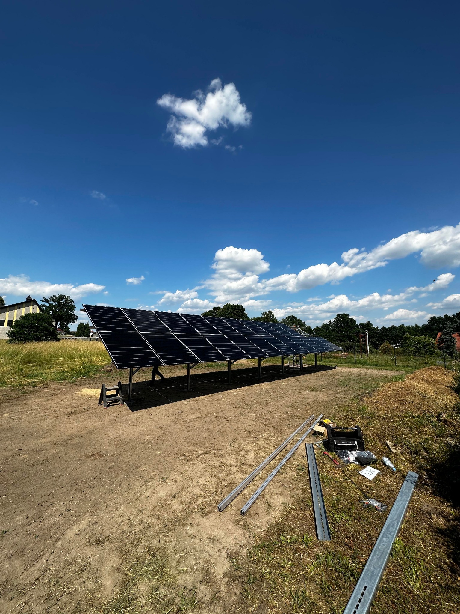 Rozległa instalacja fotowoltaiczna na metalowej konstrukcji w trakcie montażu, widoczne narzędzia i elementy montażowe na ziemi, błękitne niebo z chmurami w tle.