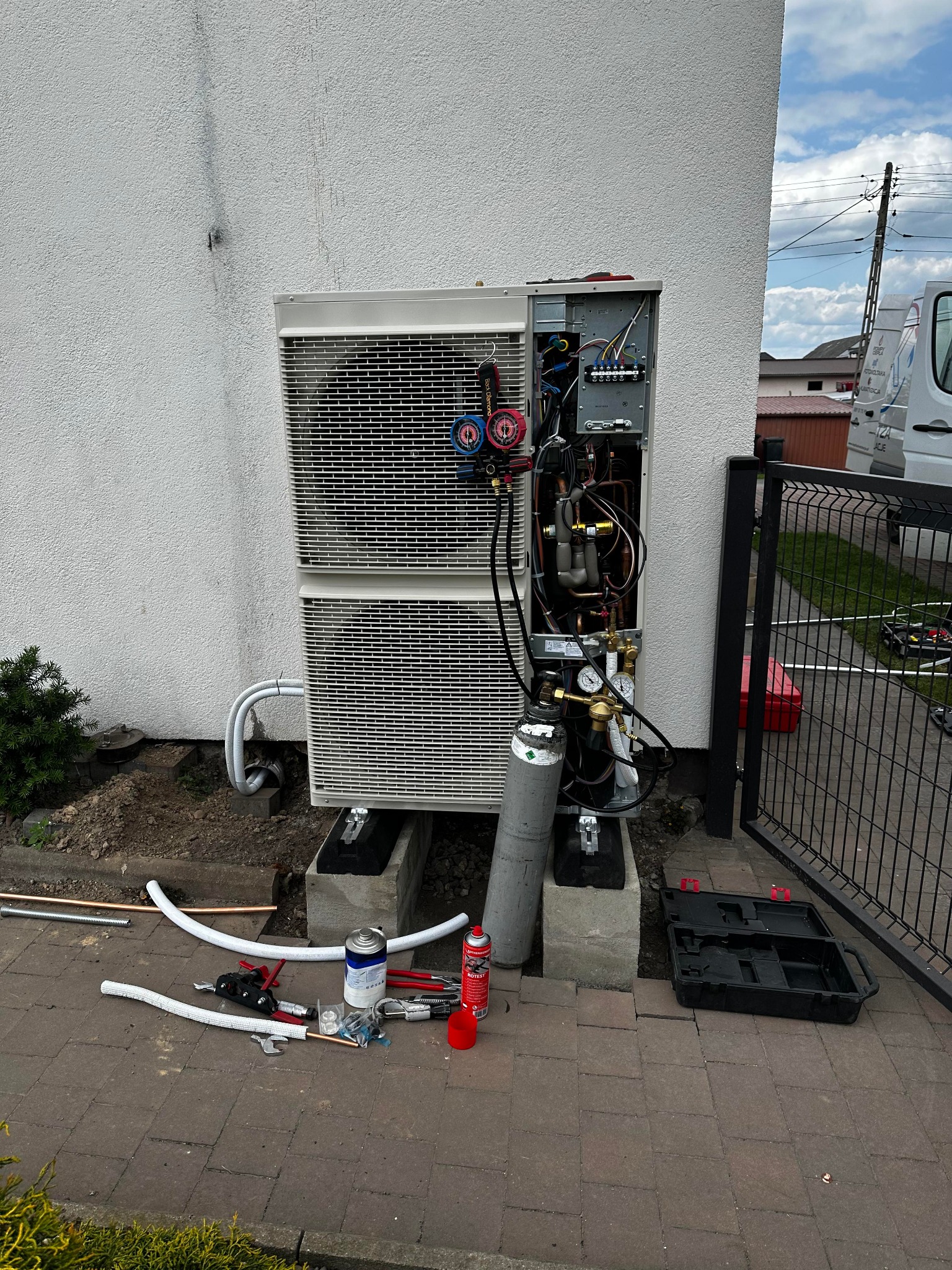 Montaż zewnętrznej jednostki pompy ciepła z otwartą obudową, widoczne elementy instalacji, butla z gazem chłodniczym, narzędzia na kostce brukowej, budynek w tle.
