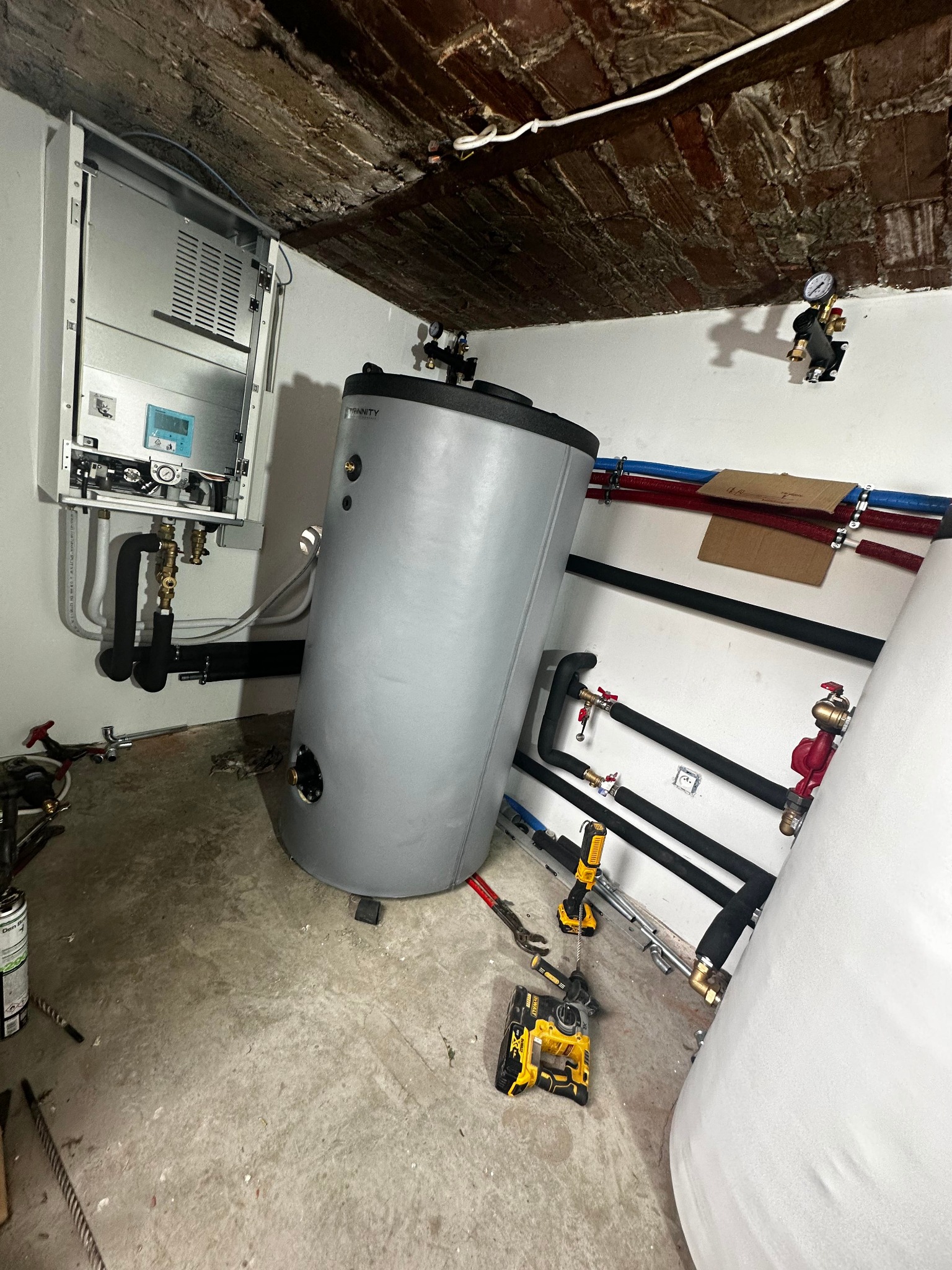 Montaż kotła grzewczego i zasobnika ciepłej wody w piwnicy z ceglanym sufitem, widoczne narzędzia DeWalt i rury instalacyjne w izolacji.