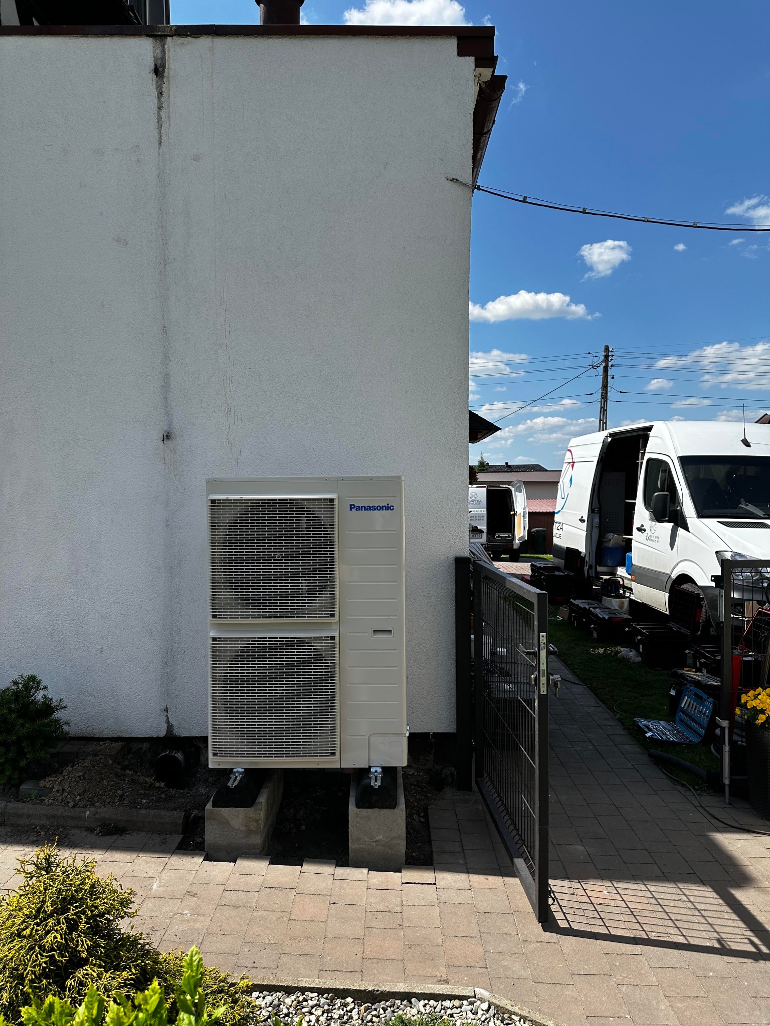 Zewnętrzna jednostka pompy ciepła Panasonic zamontowana przy białej ścianie domu, widoczne dwa samochody serwisowe firmy w tle, brama wejściowa i narzędzia.