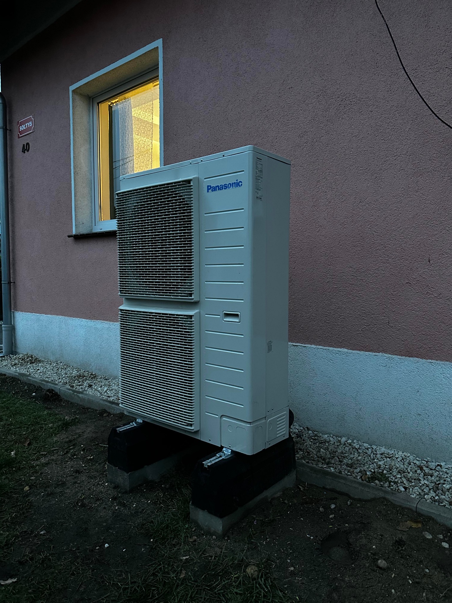 Zewnętrzna jednostka pompy ciepła Panasonic zamontowana na betonowych podstawach przy ścianie budynku z oknem, widoczne elementy instalacji.