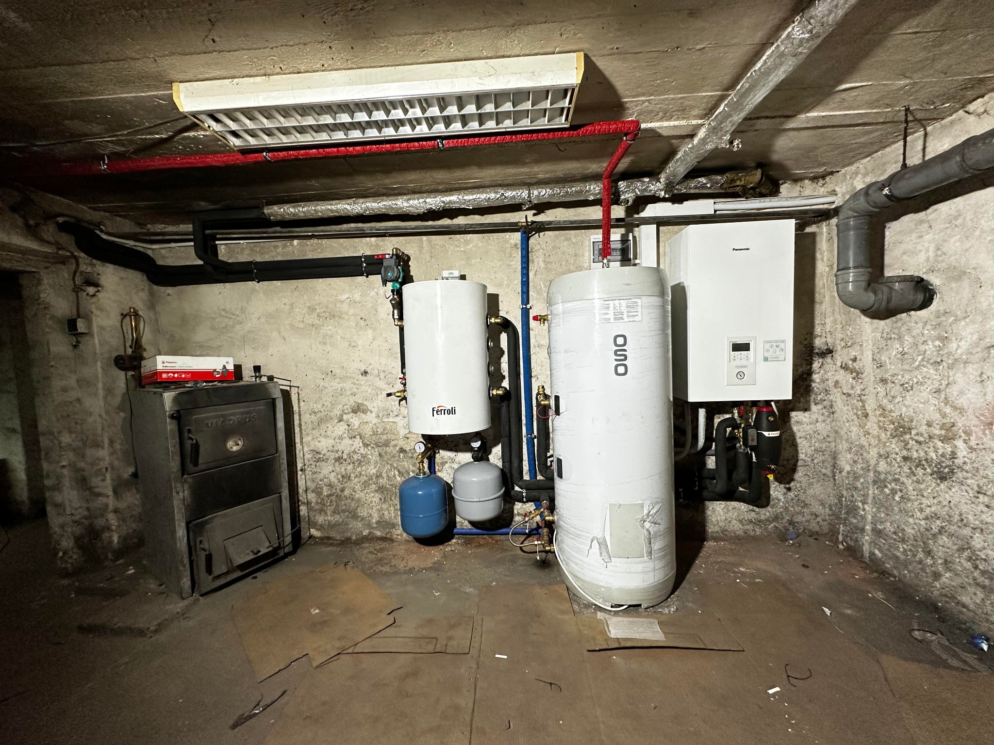 Piwnica z zainstalowanym kotłem Viadrus, dwoma zbiornikami Ferroli i OSO oraz wiszącym kotłem gazowym Panasonic, rury w izolacji czerwonej, podłoga wyłożona kartonem.