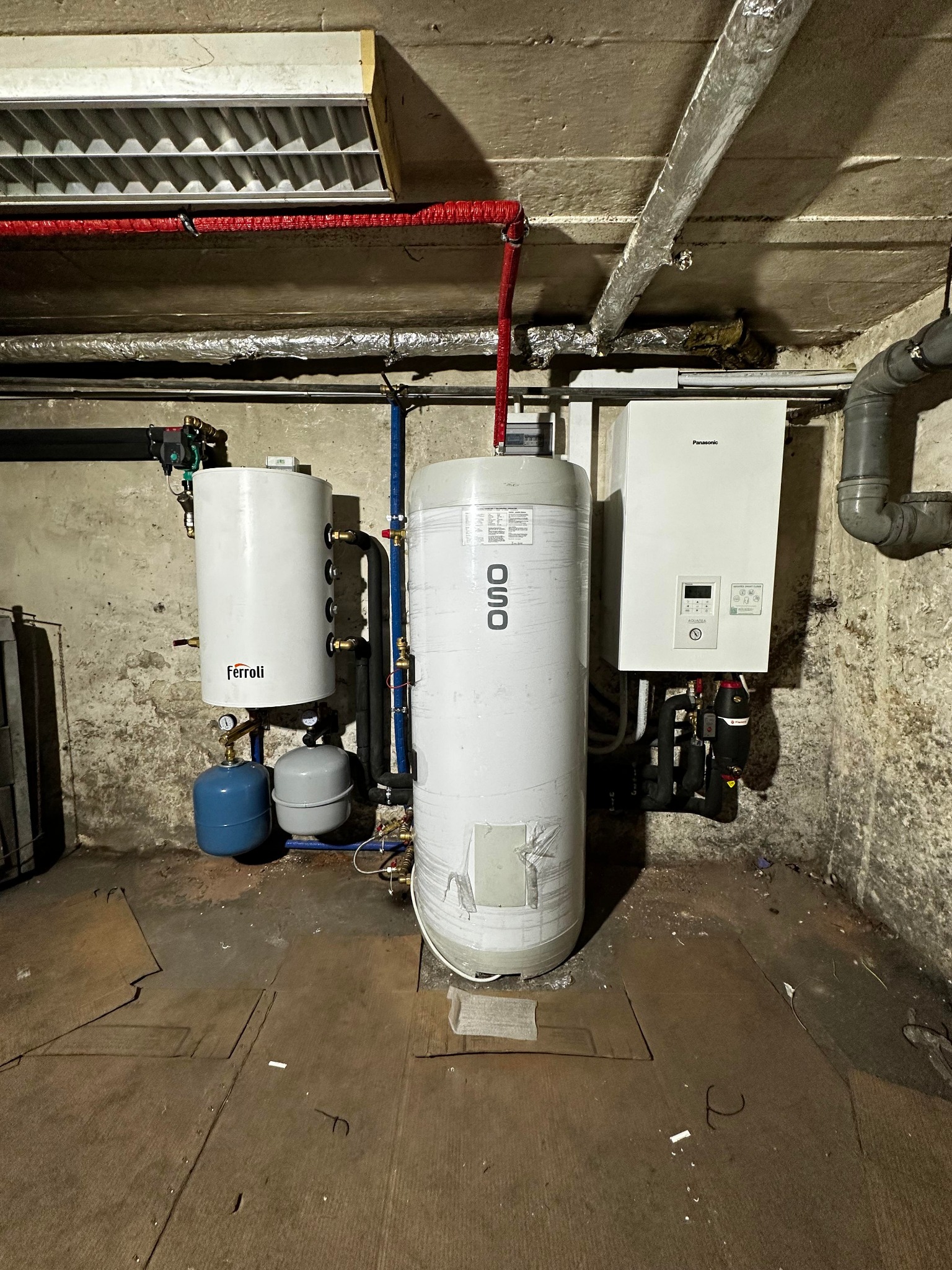 Kotłownia w piwnicy: biały piec gazowy Panasonic, dwa białe zbiorniki Ferolli i OSO, rury w izolacji, betonowy sufit i podłoga wyłożona płytami.