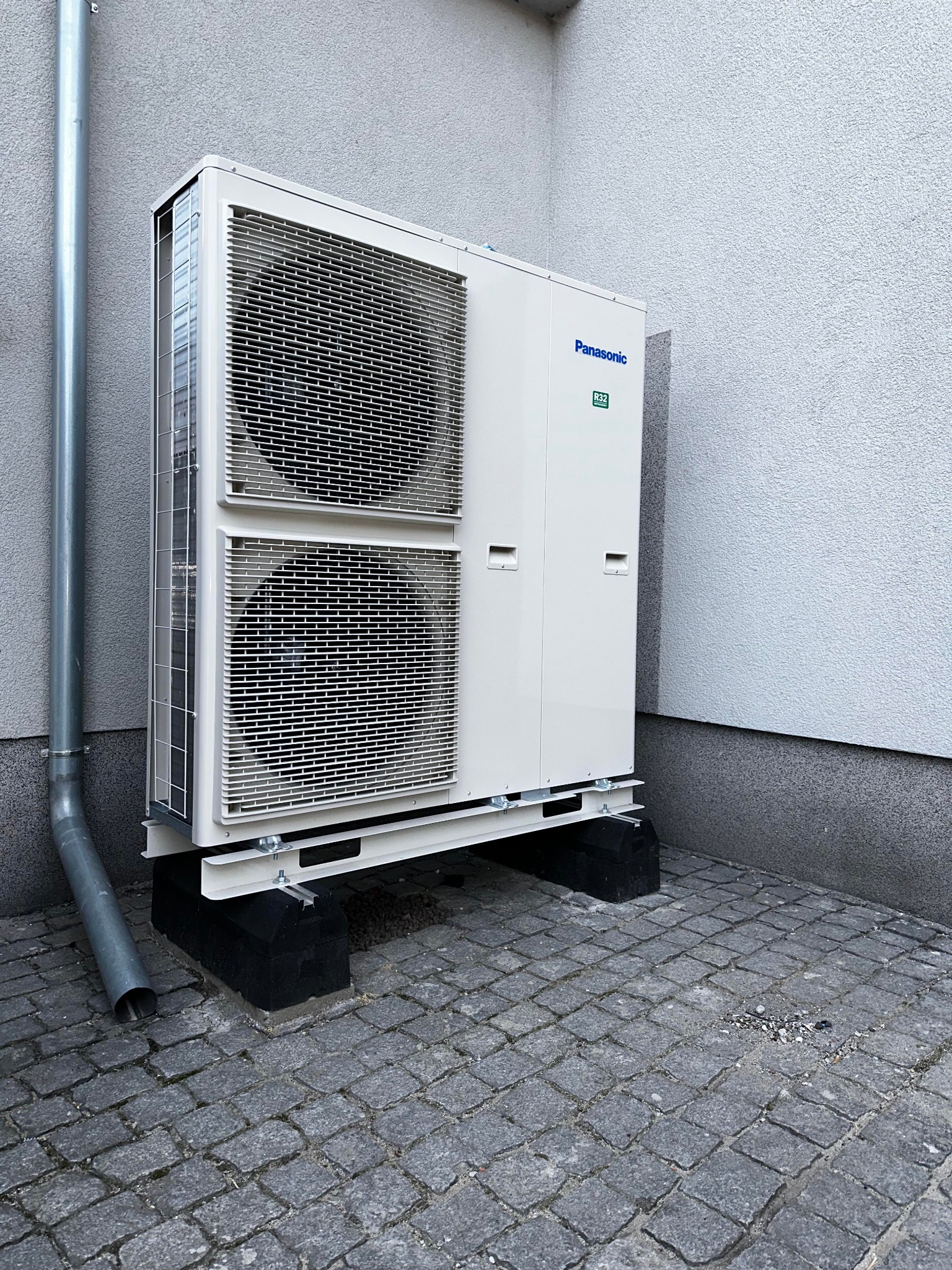 Zewnętrzna jednostka pompy ciepła Panasonic R32, zamontowana na czarnych podstawach z szarej kostki brukowej, widoczny fragment rury spustowej przy ścianie budynku z szarym tynkiem.