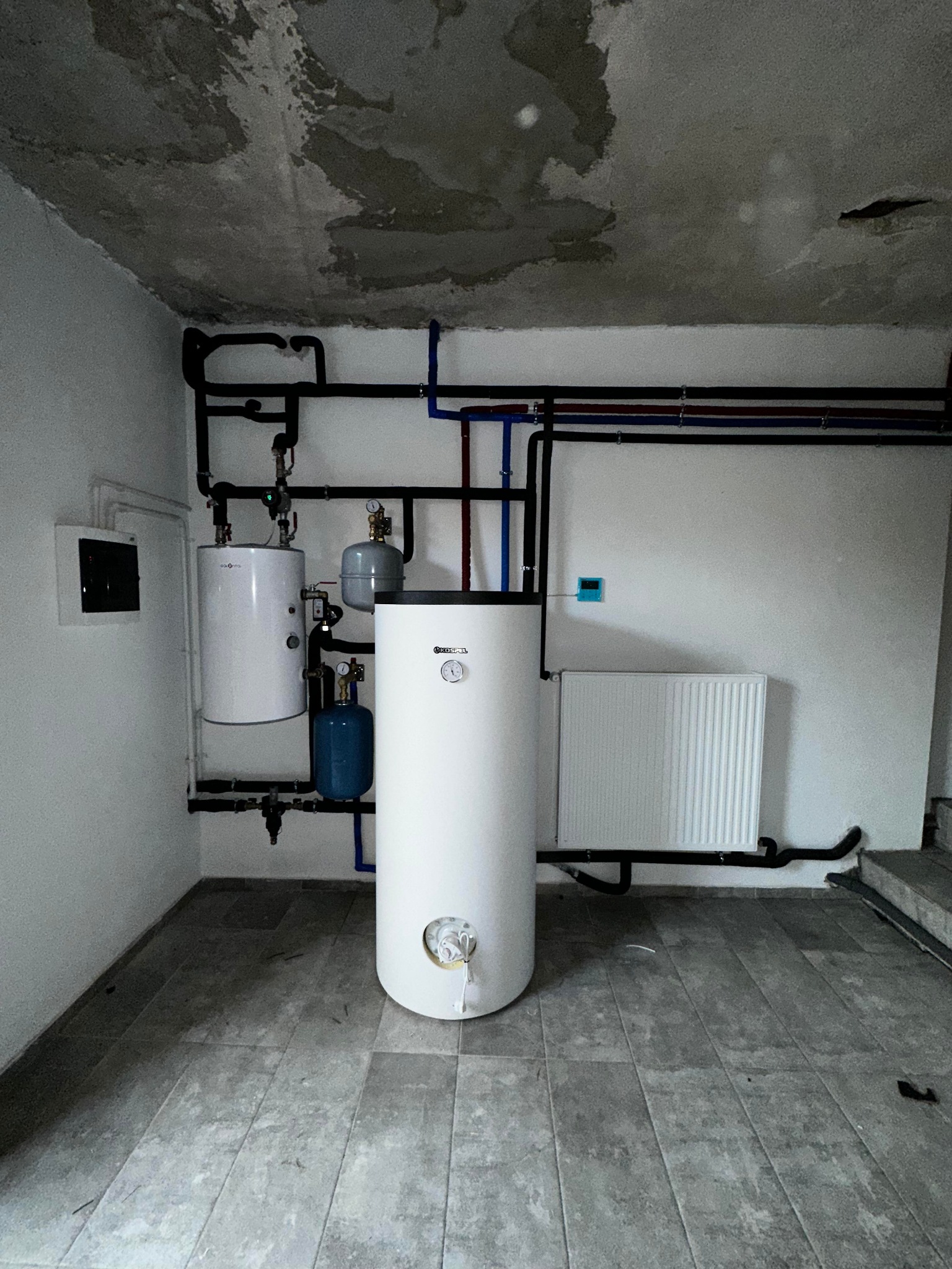 Nowoczesna kotłownia z białymi zbiornikami, licznymi rurami w kolorze czarnym i niebieskim, grzejnikiem panelowym i szarą podłogą z płytek, surowy sufit z widocznymi śladami wilgoci.