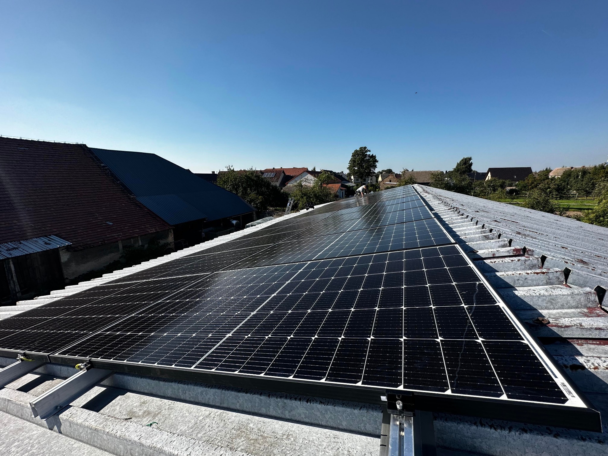 Panele fotowoltaiczne zamontowane na blaszanym dachu w wiejskiej scenerii, widoczne domy z czerwonymi dachówkami i pracownik w oddali.