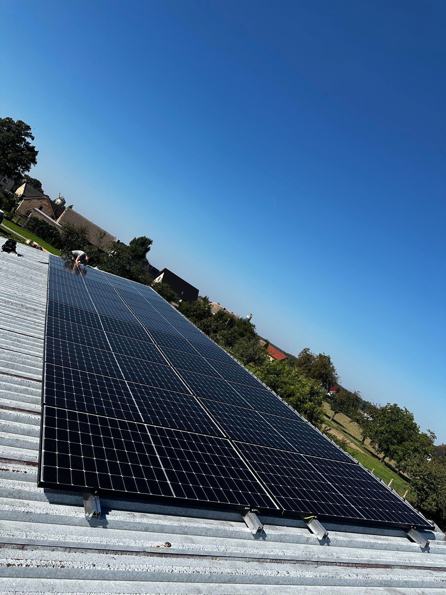 Instalacja paneli słonecznych na blaszanym dachu budynku w słoneczny dzień, widok z dołu pod kątem, pracownik w oddali przy panelach.