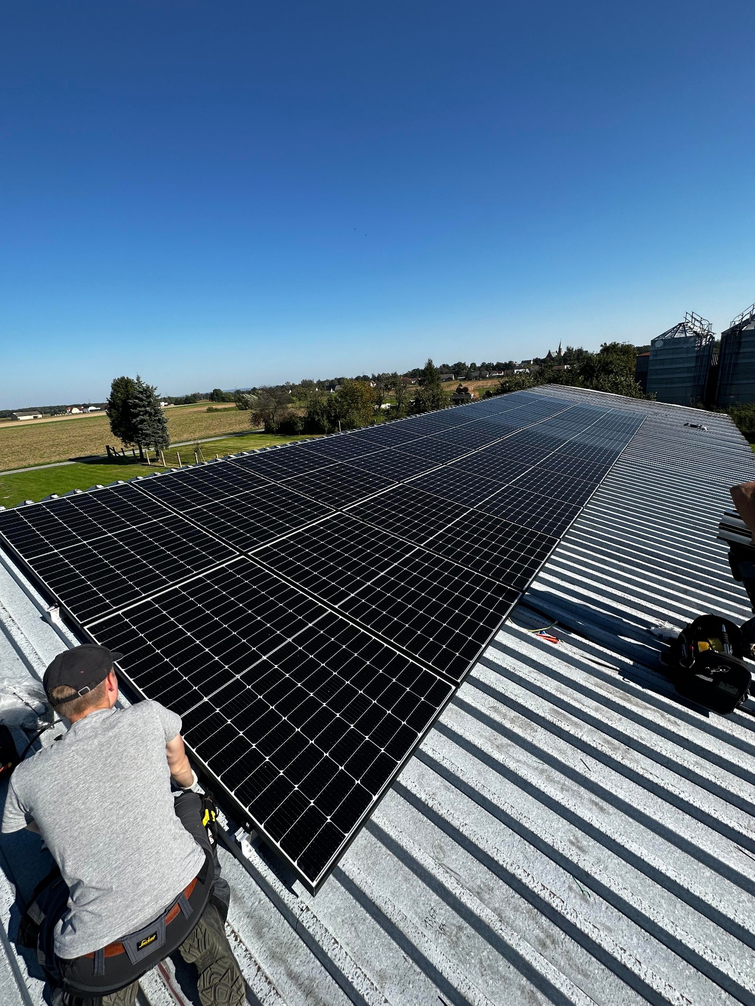 Monter w szarej koszulce instaluje panele słoneczne na blaszanym dachu; w tle wieś i silosy zbożowe pod błękitnym niebem.