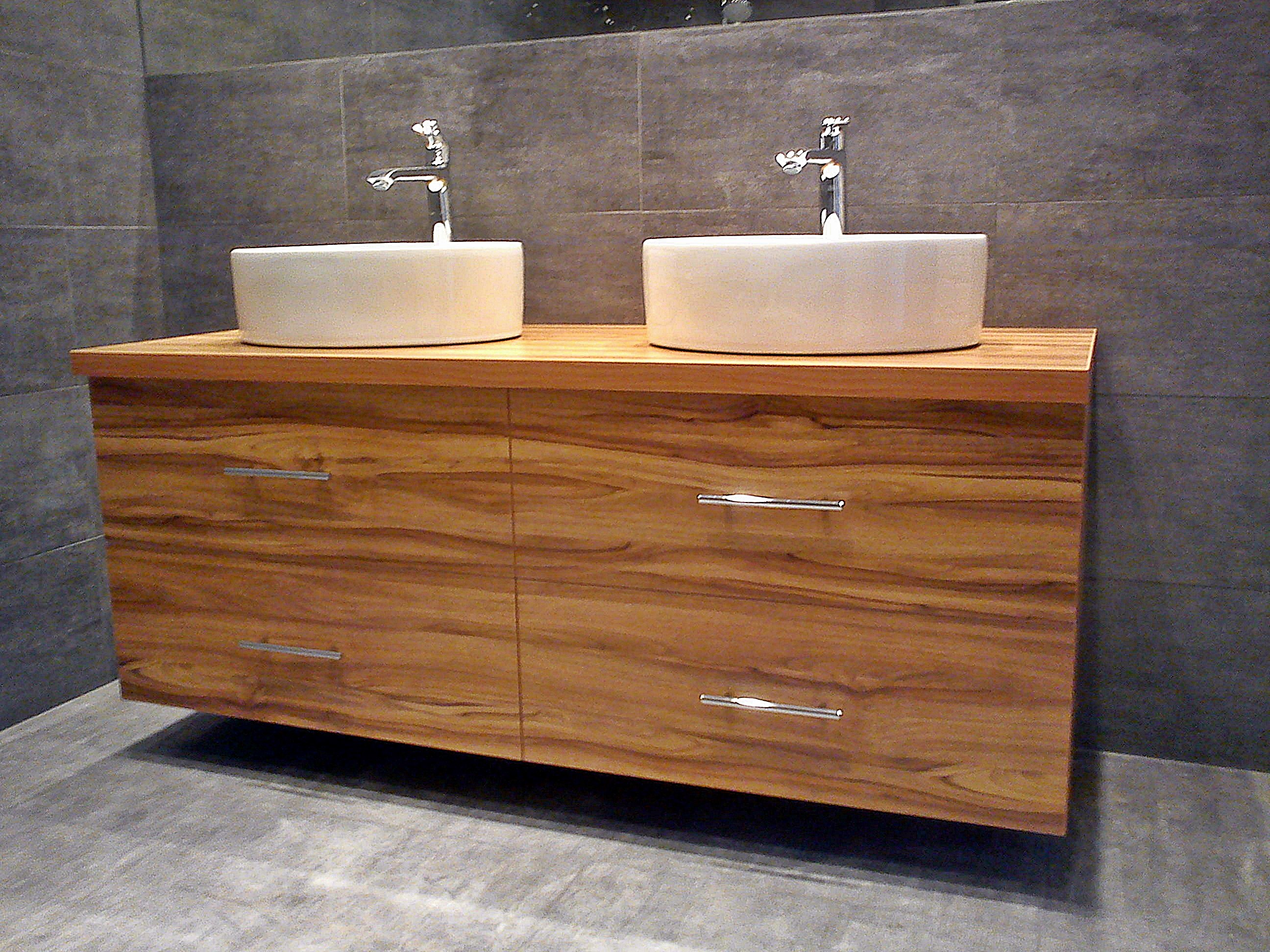 Nowoczesna szafka łazienkowa z drewna o wyraźnym usłojeniu, z dwoma okrągłymi umywalkami nablatowymi i chromowanymi bateriami, na tle szarej ściany wyłożonej płytkami.
