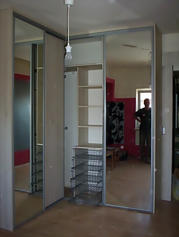 Wnętrze z otwartą szafą z przesuwnymi, lustrzanymi drzwiami, widoczne półki i wysuwane kosze druciane, w tle odbicie osoby w pokoju z czerwonym akcentem na ścianie.