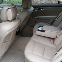 Wnętrze luksusowego samochodu z beżową skórzaną tapicerką, widok na tylne siedzenia i podłokietnik, drewniane wykończenia na drzwiach, dywaniki podłogowe.