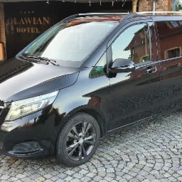 Czarny Mercedes-Benz Vito zaparkowany przed hotelem Oławian, z bryczką ozdobną w tle.