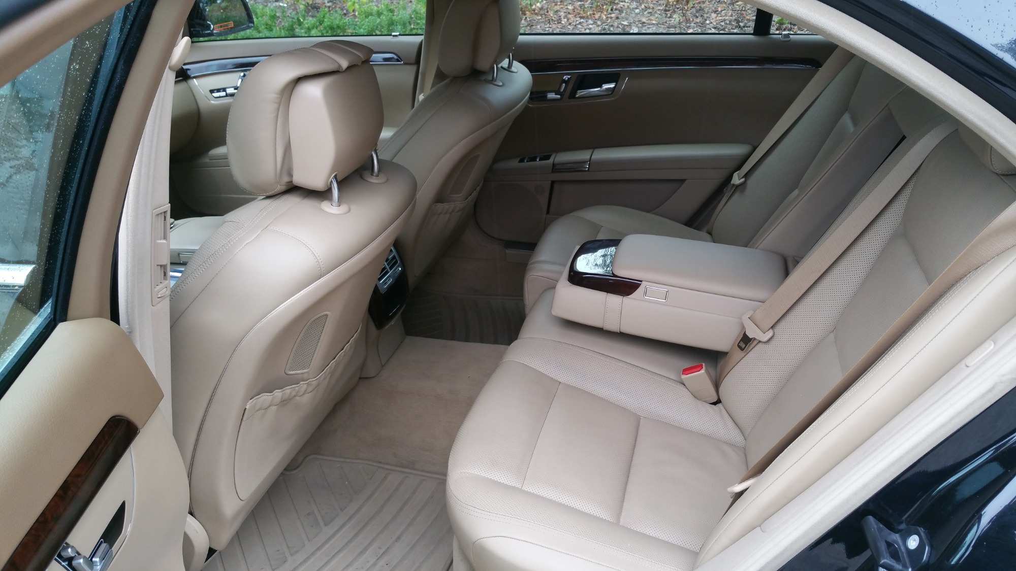 Wnętrze luksusowego samochodu z beżową skórzaną tapicerką, widok na tylne siedzenia i podłokietnik, drewniane wykończenia na drzwiach, dywaniki podłogowe.