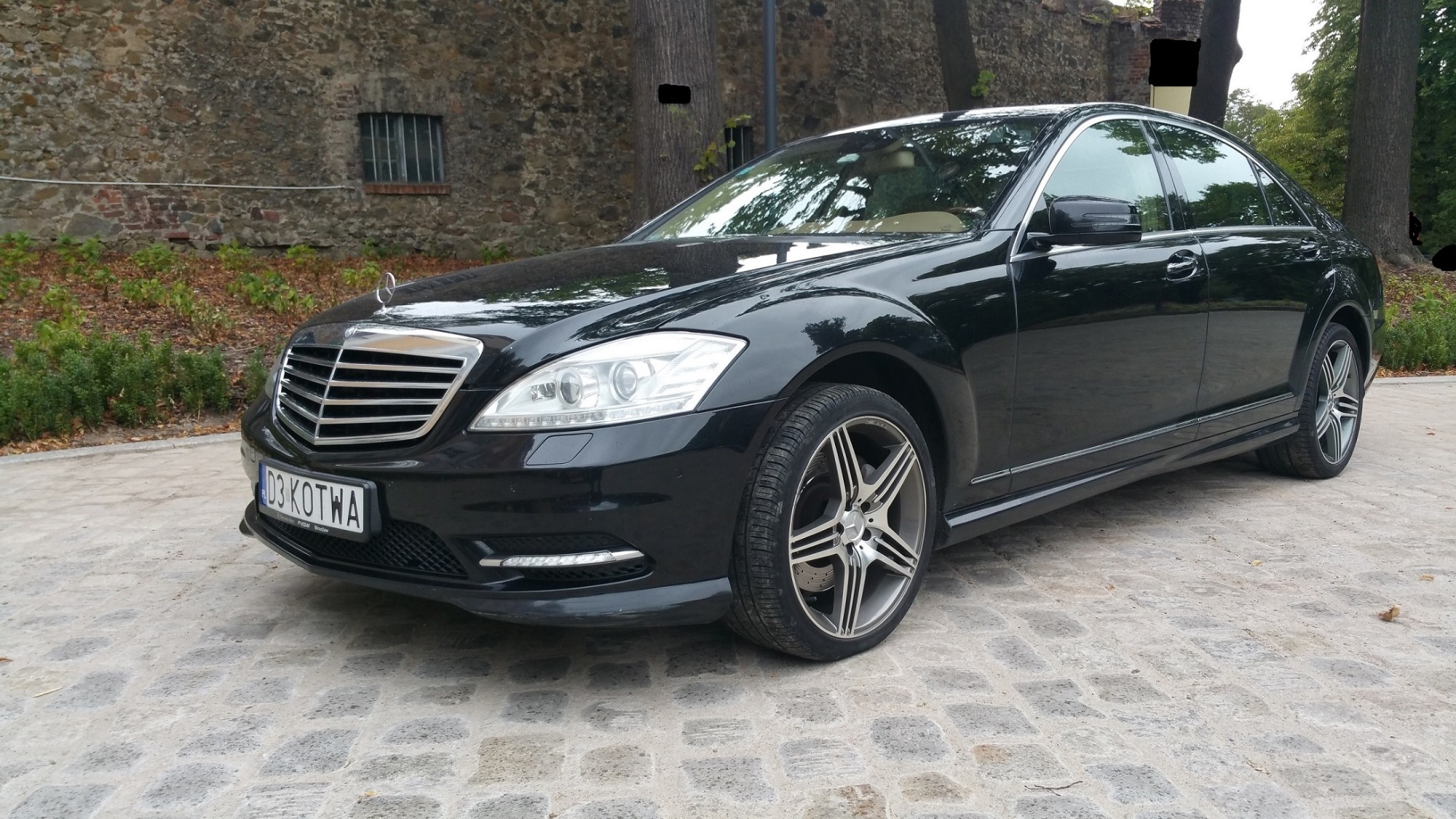 Czarna limuzyna Mercedes-Benz klasy S z chromowanymi elementami, zaparkowana na brukowanej nawierzchni z kamiennym murem i drzewami w tle.