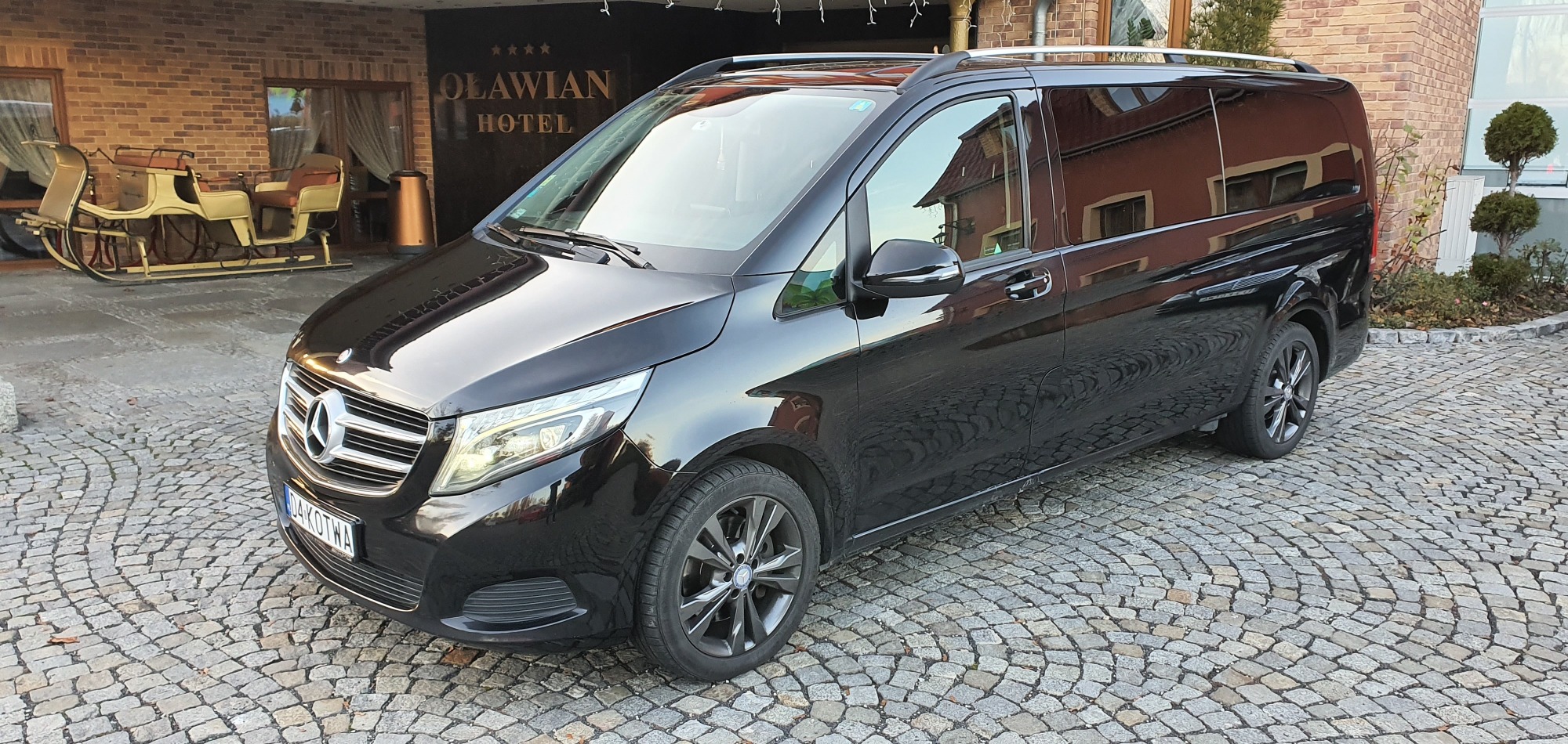 Czarny Mercedes-Benz Vito zaparkowany przed hotelem Oławian, z bryczką ozdobną w tle.