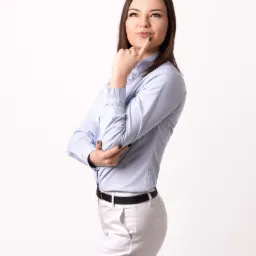 Kobieta w błękitnej koszuli w paski i białych spodniach, z palcem wskazującym przy ustach, zamyślona, patrzy w górę na białym tle.