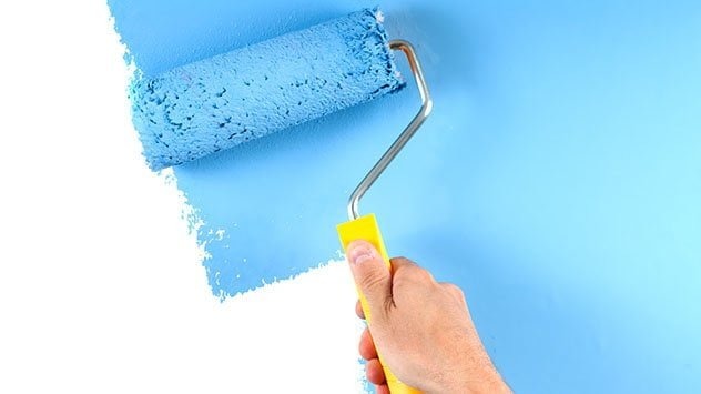 Wałek malarski z żółtą rączką w dłoni, aplikujący jasnoniebieską farbę na ścianę, kontrastując z białym fragmentem powierzchni.