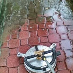 Czyszczenie czerwonej kostki brukowej za pomocą myjki ciśnieniowej z okrągłą dyszą; widoczna różnica w kolorze pomiędzy umytą a brudną powierzchnią, obok siatka ogrodzeniowa.