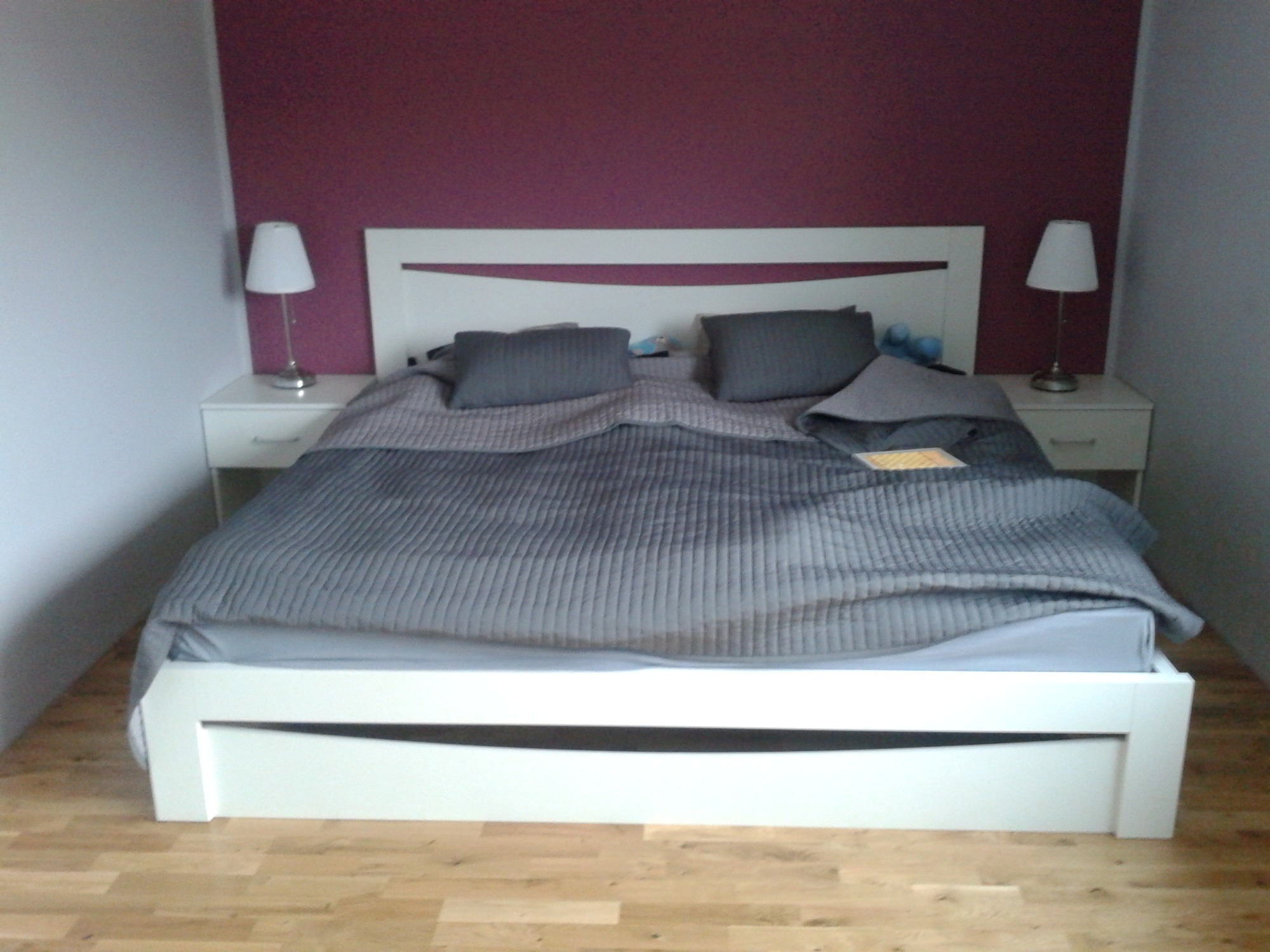 Białe łóżko z szufladami i zagłówkiem z wstawką w pokoju z fioletową ścianą, obok szafki nocne z lampkami.