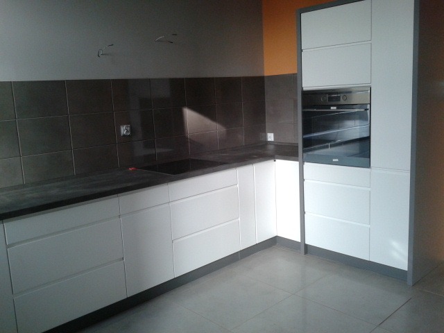 Nowoczesna kuchnia z białymi frontami szafek, ciemnym blatem i zabudowaną piekarnikiem, na tle brązowych płytek ściennych.