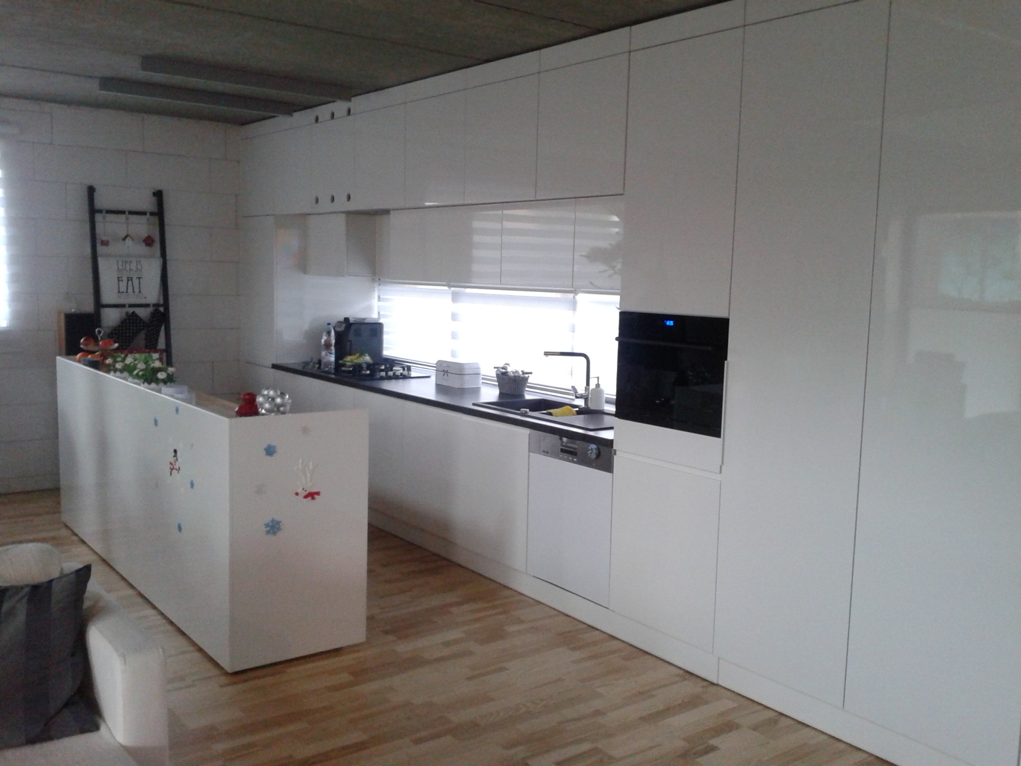 Nowoczesna kuchnia z białymi, lakierowanymi frontami, czarnym blatem i wyspą ozdobioną zimowymi motywami, widoczna z perspektywy salonu.