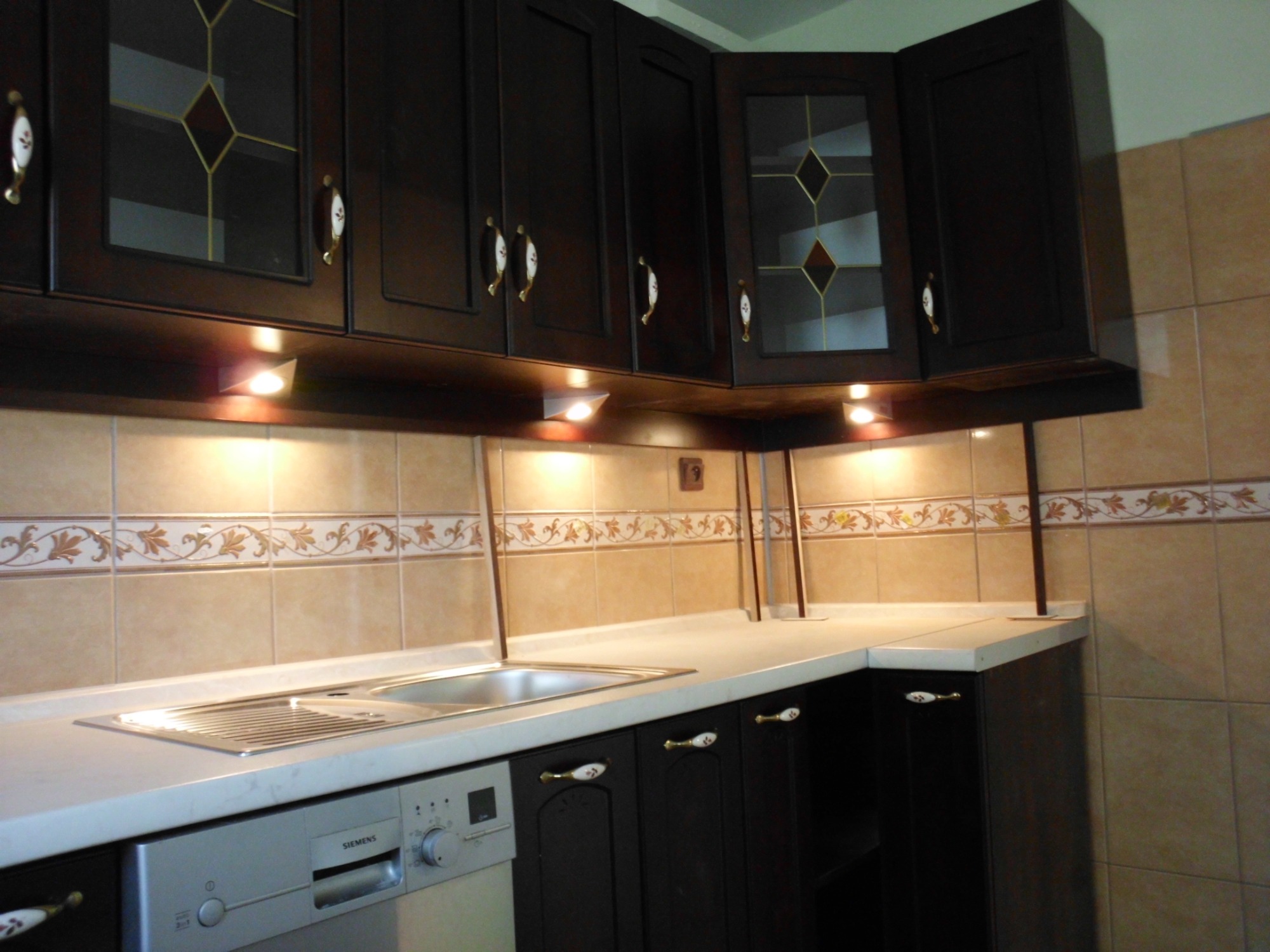 Ciemnobrązowe szafki kuchenne z przeszklonymi drzwiczkami, ozdobnymi uchwytami i podświetleniem LED nad blatem, z widocznym fragmentem zlewu i zmywarki Siemens.