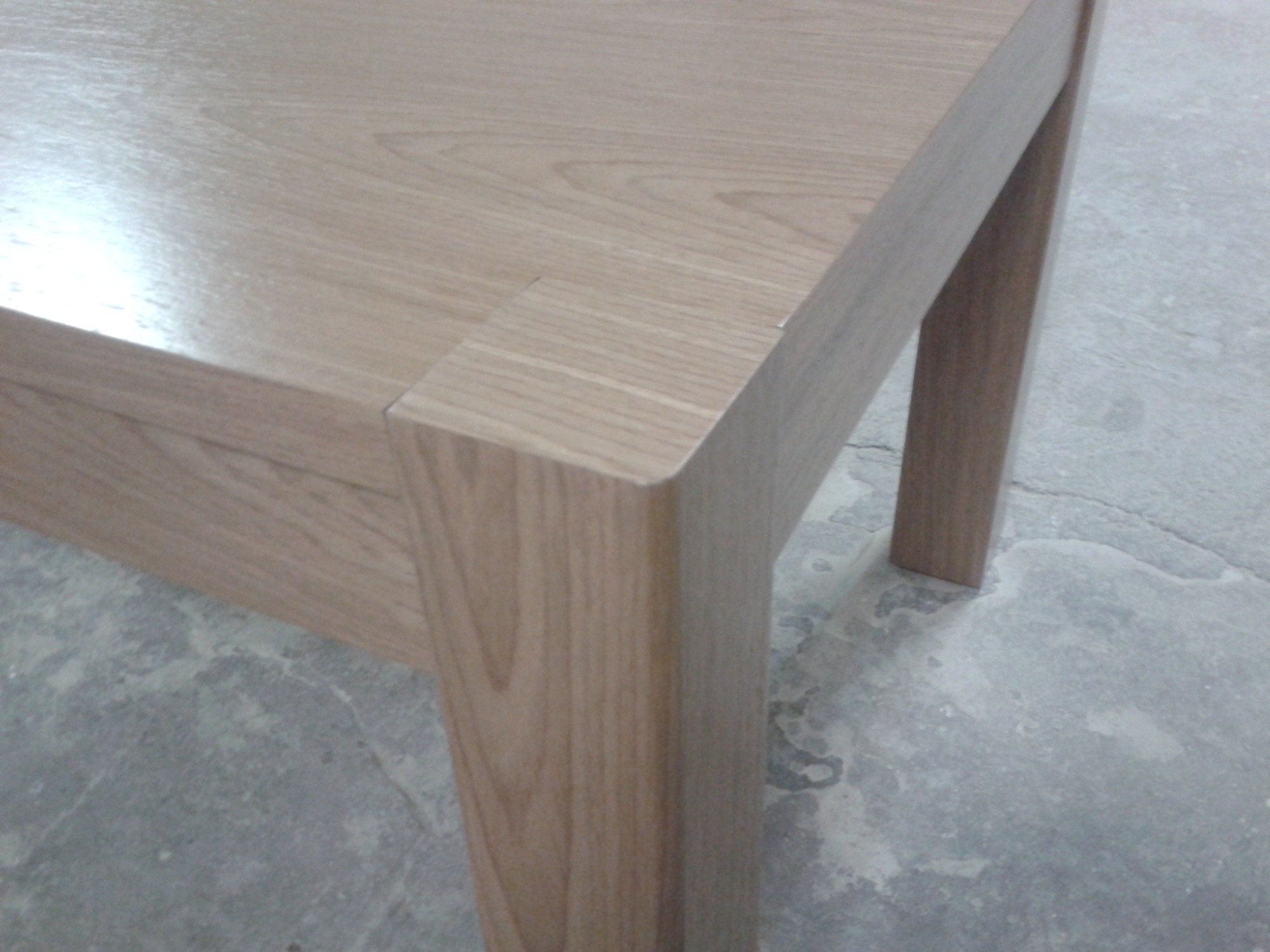 Fragment drewnianego stołu z blatem o jasnym odcieniu drewna, widoczny narożnik i noga, na tle szarej, betonowej podłogi.