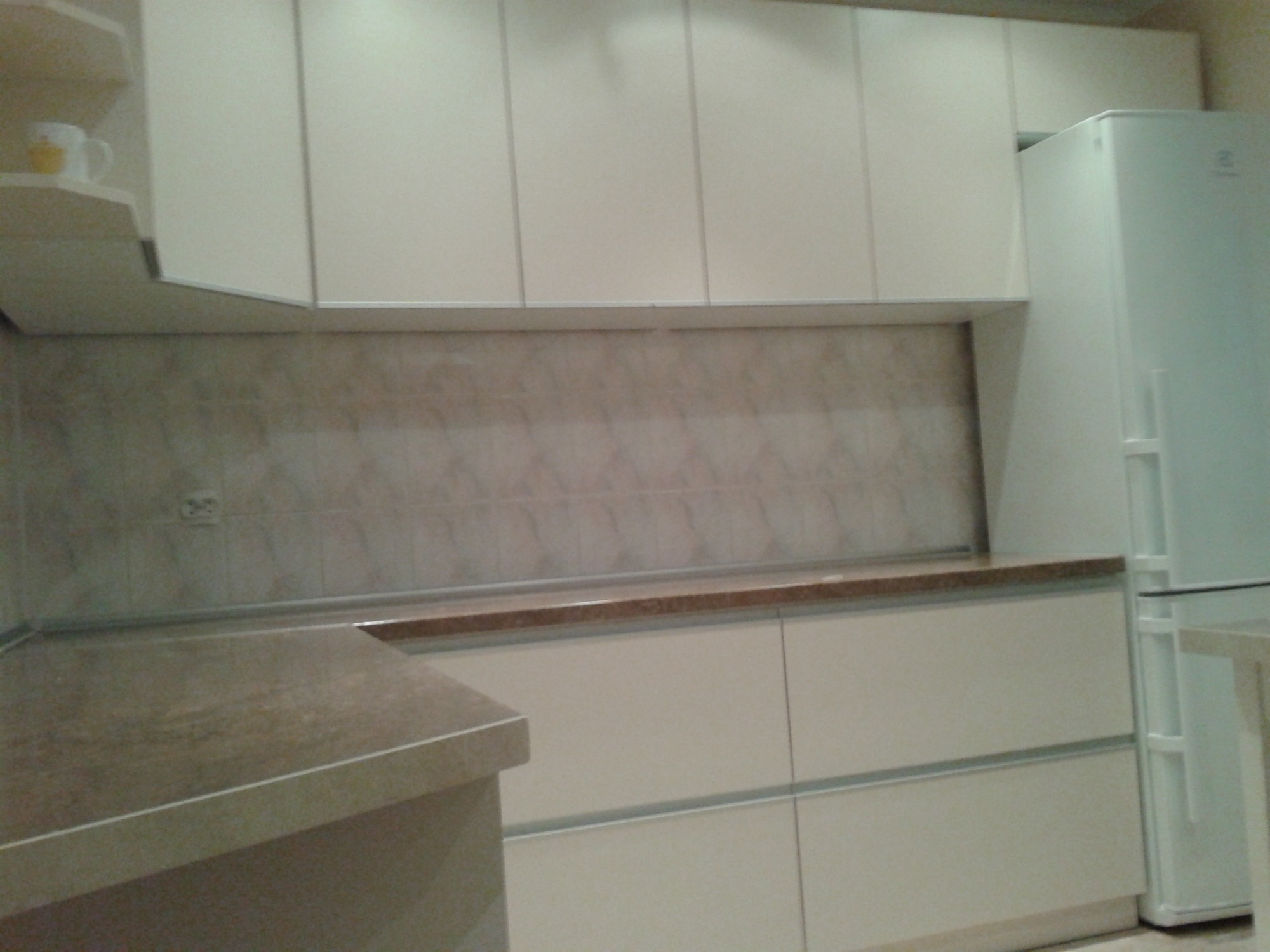 Jasna, nowoczesna zabudowa kuchenna z białymi frontami, brązowym blatem i płytkami w odcieniach beżu. W tle widoczna biała lodówka.