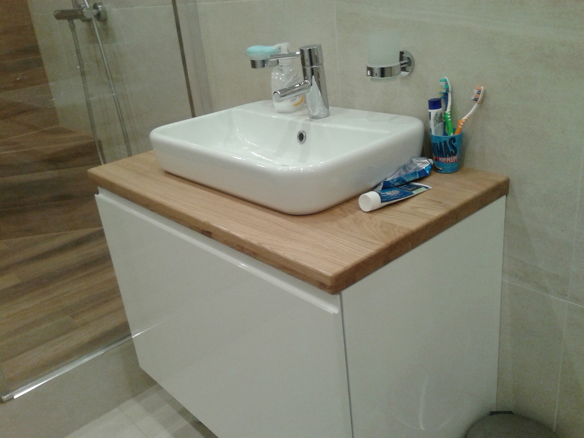 Biała umywalka nablatowa z chromowaną baterią na szafce z drewnianym blatem w łazience, obok kubek ze szczoteczkami do zębów i tubka pasty.
