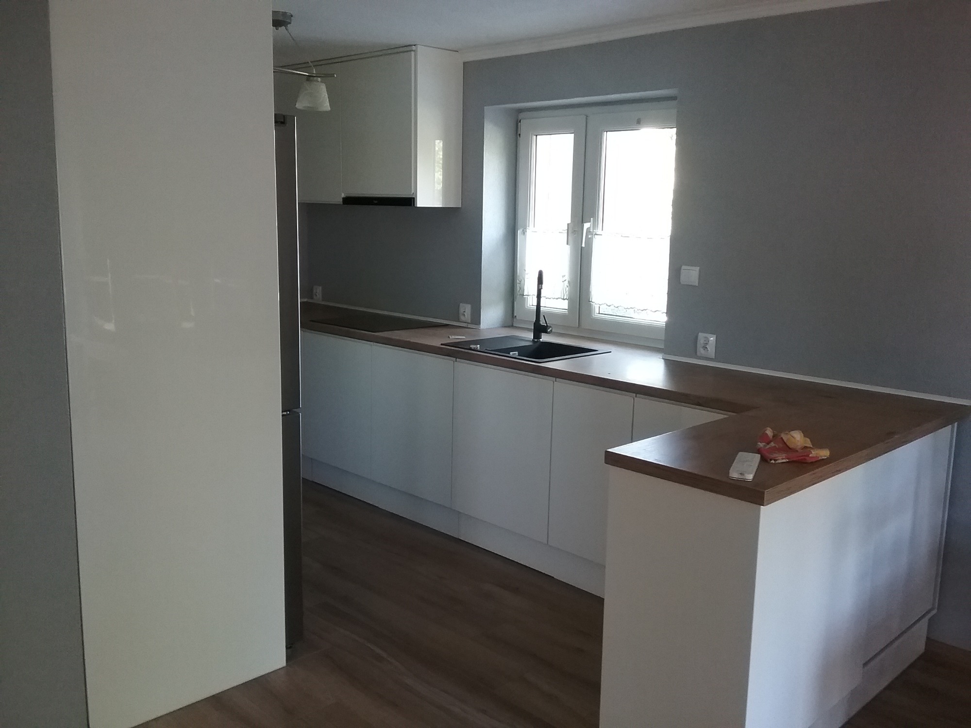 Nowoczesna kuchnia z białymi frontami, drewnianym blatem i czarnym zlewozmywakiem pod oknem, częściowo zasłonięta przez białą ścianę po lewej stronie, z pilotem i ściereczką leżącymi na blacie.