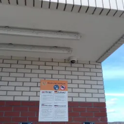 Kamera monitoringu zamontowana pod sufitem nad wejściem do budynku z ceglaną elewacją, widoczne oświetlenie i tabliczka informacyjna.