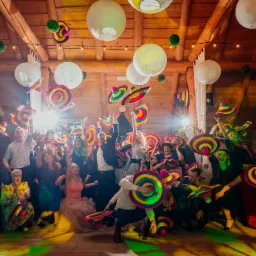 Grupa ludzi wewnątrz drewnianego budynku, trzymająca w górze kolorowe sombrera, pod dekoracyjnymi lampionami i oświetleniem, podczas imprezy.