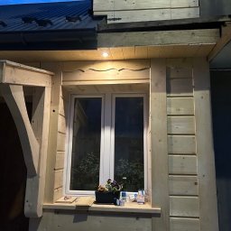 T&M BUDOWNICTWO SPÓŁKA Z OGRANICZONĄ ODPOWIEDZIALNOŚCIĄ - Fragment elewacji drewnianego domu z oknem, ozdobnymi wykończeniami i daszkiem; na parapecie doniczka z kwiatami i opakowania leków.