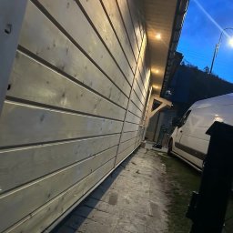 T&M BUDOWNICTWO SPÓŁKA Z OGRANICZONĄ ODPOWIEDZIALNOŚCIĄ - Elewacja budynku z drewnianych paneli pomalowanych na jasny kolor, widok z dołu wzdłuż ściany, z widocznymi punktami świetlnymi pod dachem i fragmentem zaparkowanego samochodu dostawczego