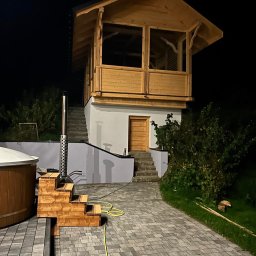 T&M BUDOWNICTWO SPÓŁKA Z OGRANICZONĄ ODPOWIEDZIALNOŚCIĄ - Dwukondygnacyjny budynek z drewna z przeszkloną górną kondygnacją, osadzony na murowanym fundamencie z zewnętrznymi schodami, obok drewniana balia z piecem i kominem, nocne oświetlenie.