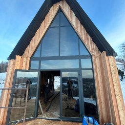 T&M BUDOWNICTWO SPÓŁKA Z OGRANICZONĄ ODPOWIEDZIALNOŚCIĄ - Surowy stan domu w kształcie litery A z drewnianą elewacją, dużym trójkątnym oknem i otwartymi szklanymi drzwiami, widoczne narzędzia i materiały budowlane na drewnianym tarasie, zimowy krajobraz...