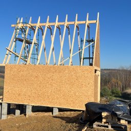 T&M BUDOWNICTWO SPÓŁKA Z OGRANICZONĄ ODPOWIEDZIALNOŚCIĄ - Wznoszenie konstrukcji domu szkieletowego na fundamencie z bloczków betonowych, widoczna drewniana więźba dachowa i płyty OSB, w tle pagórkowaty krajobraz.