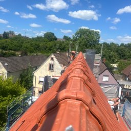 T&M BUDOWNICTWO SPÓŁKA Z OGRANICZONĄ ODPOWIEDZIALNOŚCIĄ - Czerwony dach z dachówki ceramicznej w trakcie krycia, widok z góry, pracownicy na rusztowaniu, błękitne niebo z chmurami w tle. Układanie dachówki w słoneczny dzień.