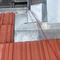 T&M BUDOWNICTWO SPÓŁKA Z OGRANICZONĄ ODPOWIEDZIALNOŚCIĄ - Detal dachu w trakcie krycia: widoczne dachówki w kolorze ceglastym, obróbki blacharskie i fragment rusztowania. Czerwony przewód na powierzchni dachu.