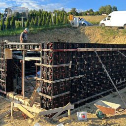 T&M BUDOWNICTWO SPÓŁKA Z OGRANICZONĄ ODPOWIEDZIALNOŚCIĄ - Szczegółowe ujęcie szalunku fundamentów z widocznymi elementami konstrukcyjnymi, wspartego na ziemi, z pracownikiem w tle i krajobrazem w tle.