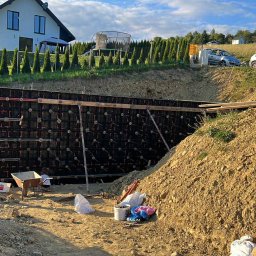 T&M BUDOWNICTWO SPÓŁKA Z OGRANICZONĄ ODPOWIEDZIALNOŚCIĄ - Wykop z szalunkiem pod fundament domu, widoczni pracownicy budowlani i sprzęt. W tle dom jednorodzinny i zieleń.