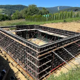 Szczegółowe ujęcie szalunku fundamentu z widoczną konstrukcją wsporczą, na tle zieleni i górskiego krajobrazu w słoneczny dzień.